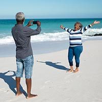 Front view of an African American couple standing on the beach with blue sky and sea in the background, the woman raising her hands and smiling, while the man takes a photo of her with his smartphone